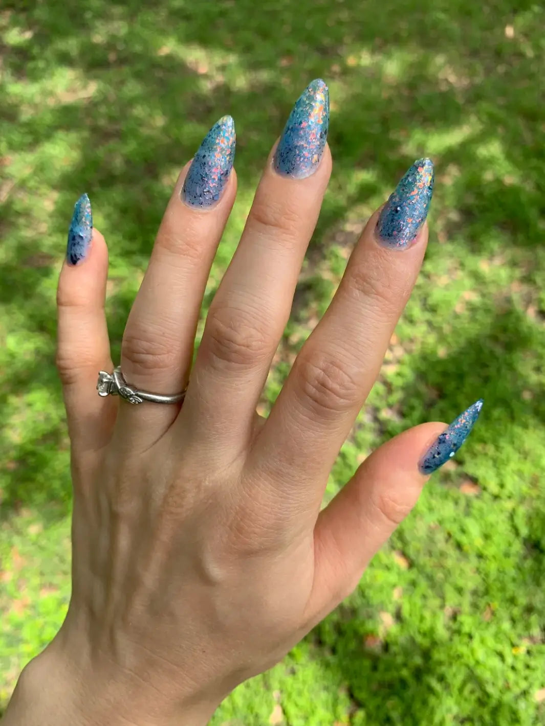 A hand against a lush green grass lawn to show off a full manicure of long, stiletto nails featuring Death Valley Nails Fish Scales Nail Polish | Twentyseven Toronto