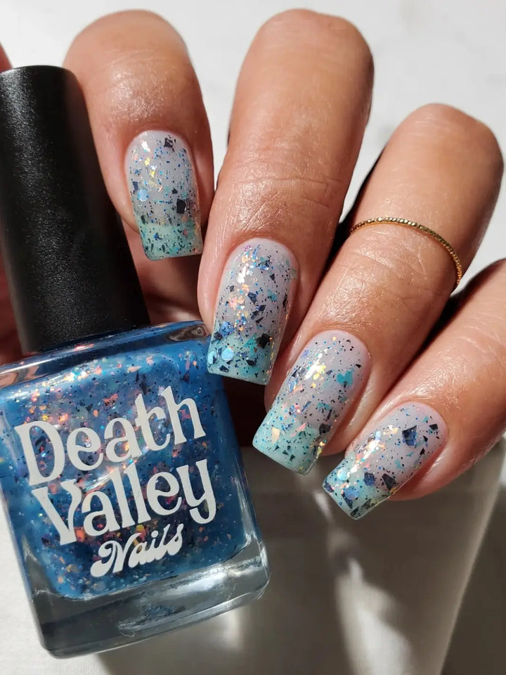 Long, square nails painted with Death Valley Nails Fish Scales Nail Polish are displayed next to the bottle, showing how the sheer blue jelly formula layers over a pale base to create a complex, shimmering finish | Twentyseven Toronto