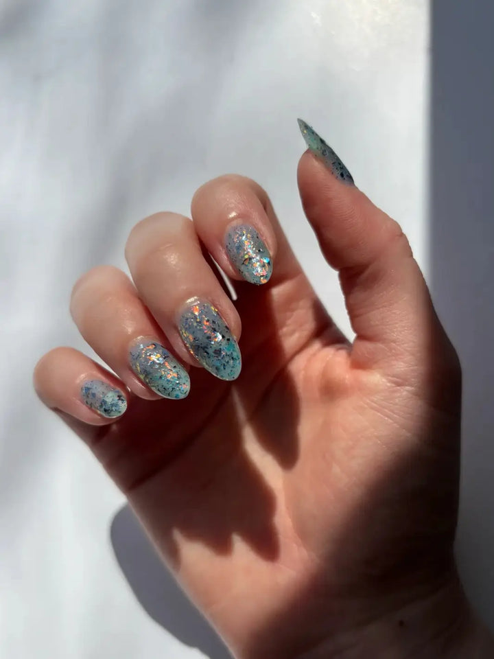 A hand with a deep skin tone and long, pointed nails displays the layered, iridescent flake effect of Death Valley Nails Fish Scales Nail Polish against a concrete background | Twentyseven Toronto