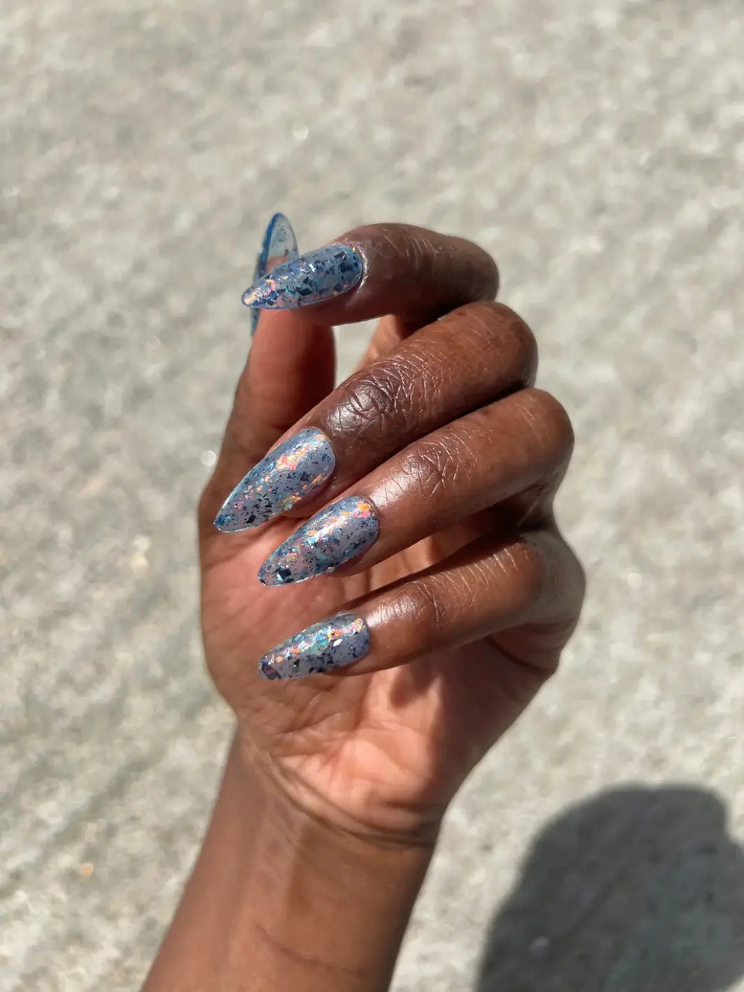 A hand with a deep skin tone holds a bottle of Death Valley Nails Fish Scales Nail Polish, matching the iridescent, speckled lacquer inside with a manicure on long, stiletto nails | Twentyseven Toronto
