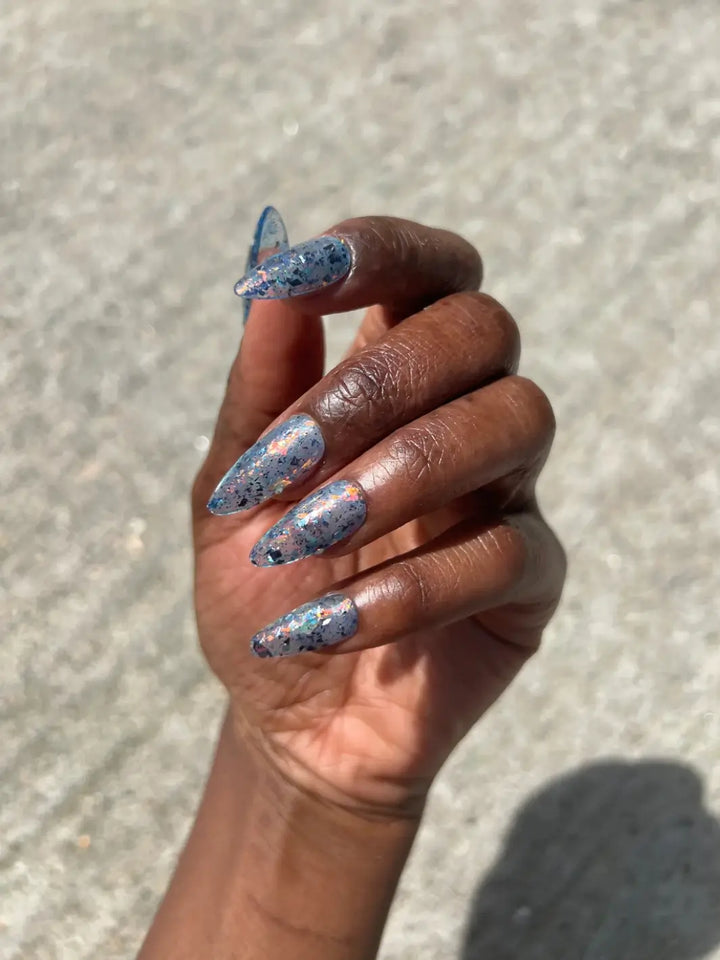A hand with a deep skin tone holds a bottle of Death Valley Nails Fish Scales Nail Polish, matching the iridescent, speckled lacquer inside with a manicure on long, stiletto nails | Twentyseven Toronto
