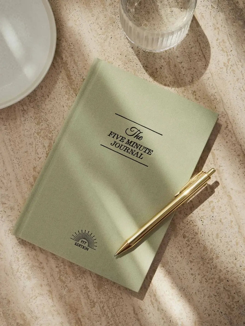 A closed, sage green hardcover of the Intelligent Change The Five Minute Journal Health & Fitness Edition lies flat on a textured stone surface next to a gold pen and a glass of water | Twentyseven Toronto