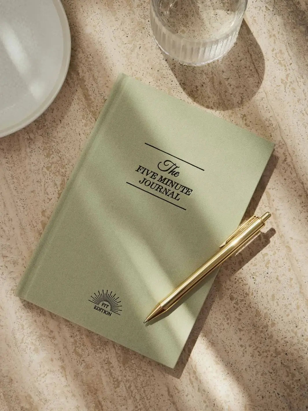 A closed, sage green hardcover of the Intelligent Change The Five Minute Journal Health & Fitness Edition lies flat on a textured stone surface next to a gold pen and a glass of water | Twentyseven Toronto
