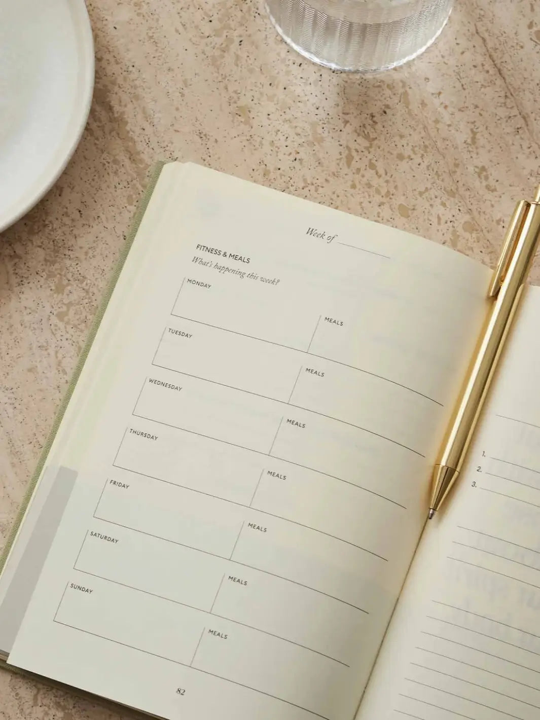 A close-up view shows the weekly fitness and meal planning grid inside the open Intelligent Change The Five Minute Journal Health & Fitness Edition with a gold pen resting on the page | Twentyseven Toronto