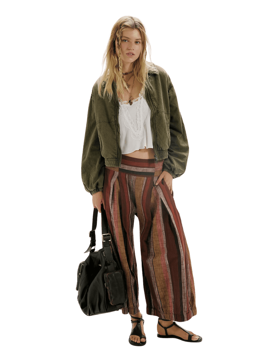 A model stands wearing a distressed olive green FP One Birdie Washed Jacket from Free People, a white cropped tank top, wide-leg striped pants in red, brown, and beige, and carrying a black duffel bag | Twentyseven Toronto