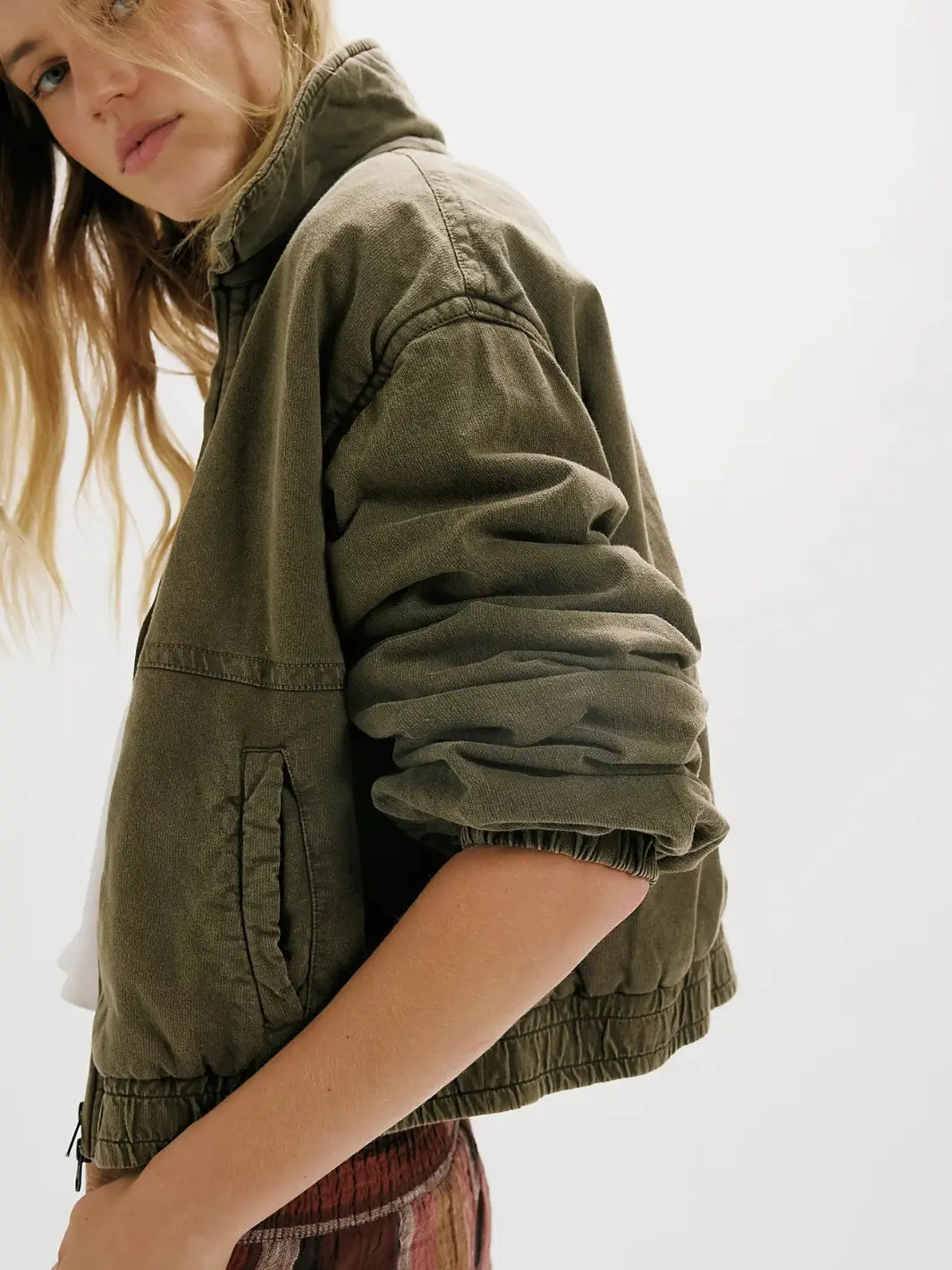 A close-up side view of a model wearing an olive green cropped utility FP One Birdie Washed Jacket from Free People with a high collar and gathered cuffs, paired with a white top and a red plaid skirt, against a bright white background | Twentyseven Toronto