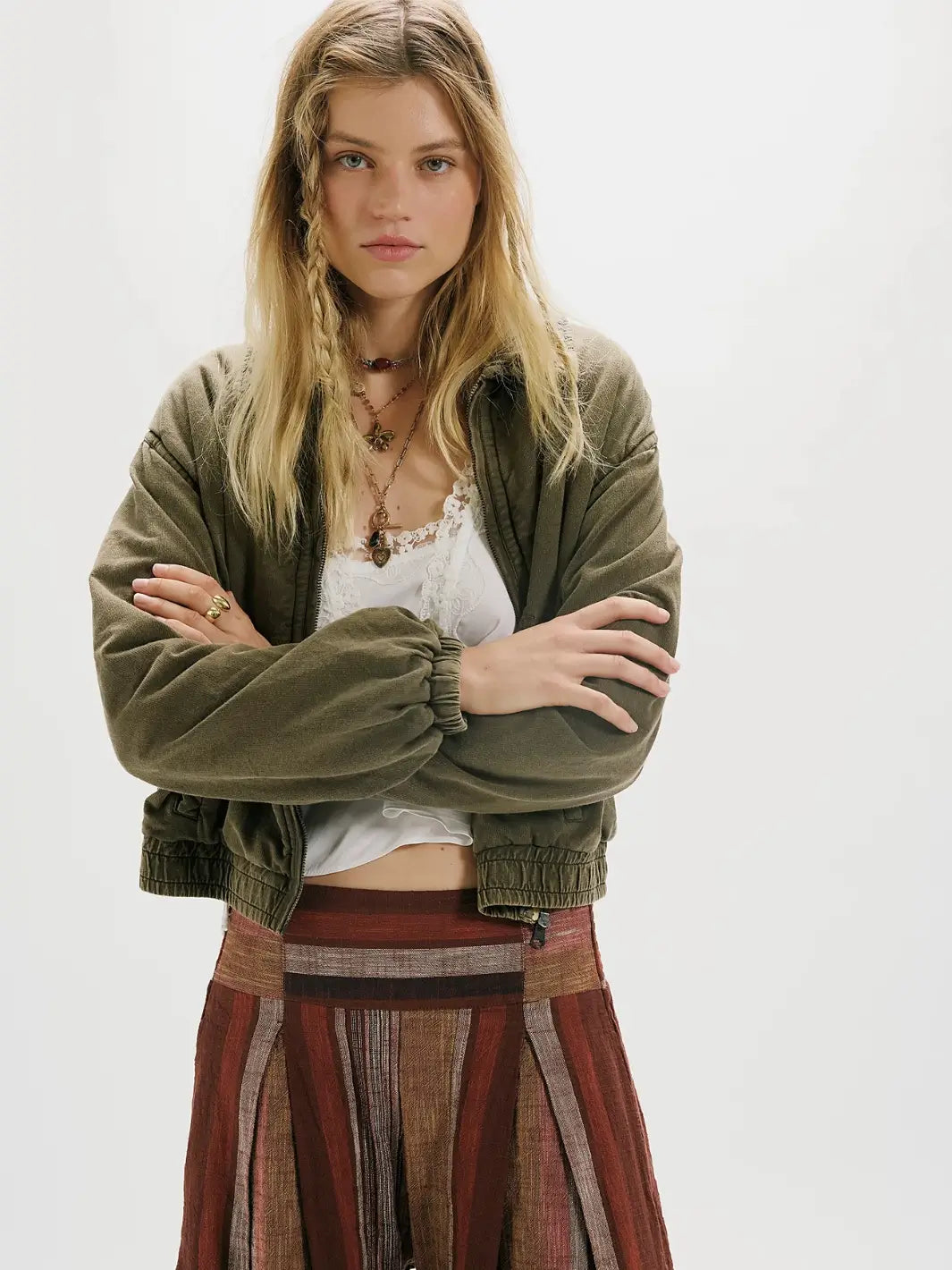 A model wearing an olive green corduroy FP One Birdie Washed Jacket from Free People over a white lace-trim top and a rust-brown striped pleated skirt, photographed against a plain white studio background | Twentyseven Toronto