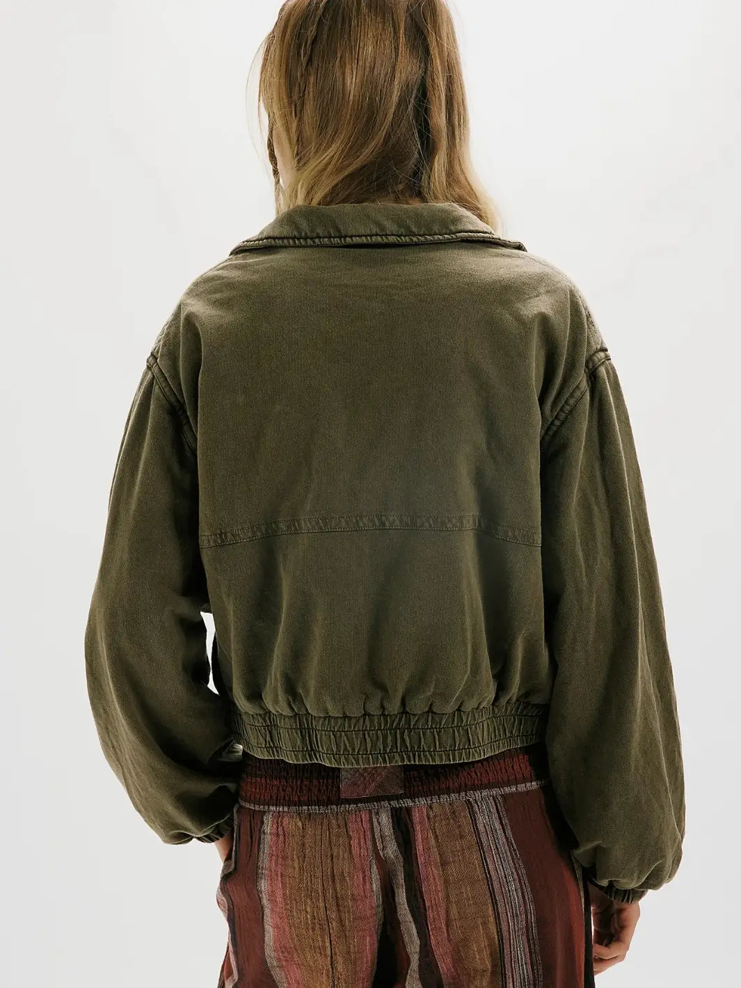A person shown from the back wearing an olive green FP One Birdie Washed Jacket with a gathered elastic hem, paired with a striped rust-brown skirt, against a plain white background | Twentyseven Toronto