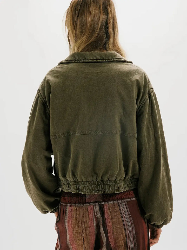 A person shown from the back wearing an olive green FP One Birdie Washed Jacket with a gathered elastic hem, paired with a striped rust-brown skirt, against a plain white background | Twentyseven Toronto