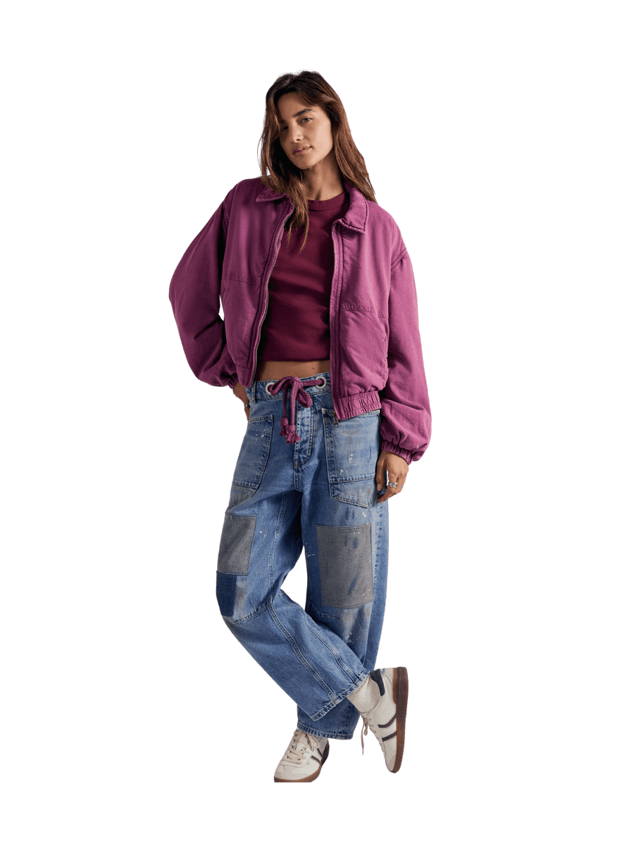 A model wearing a purple cropped bomber FP One Birdie Washed Jacket in Roan Rouge from Free People over a dark red crop top, paired with light blue patchwork denim jeans and white sneakers | Twentyseven Toronto