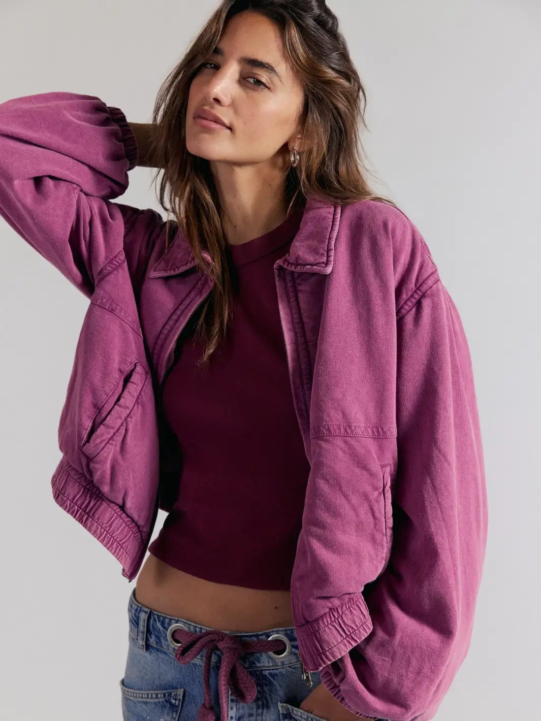 A woman wearing a dusty purple cropped FP One Birdie Washed Jacket in Roan Rouge from Free People over a matching dark plum crop top and blue jeans, photographed against a plain light gray studio background | Twentyseven Toronto