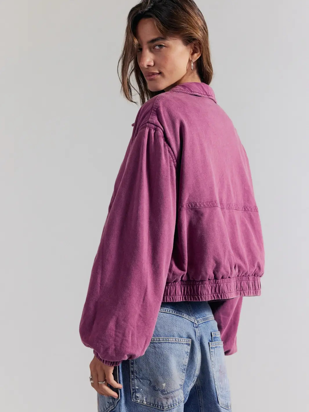 A model wearing a cropped, oversized purple FP One Birdie Washed Jacket in Roan Rouge with voluminous balloon sleeves and elasticated hem, paired with blue jeans, shown from a three-quarters back view against a light gray studio background | Twentyseven Toronto