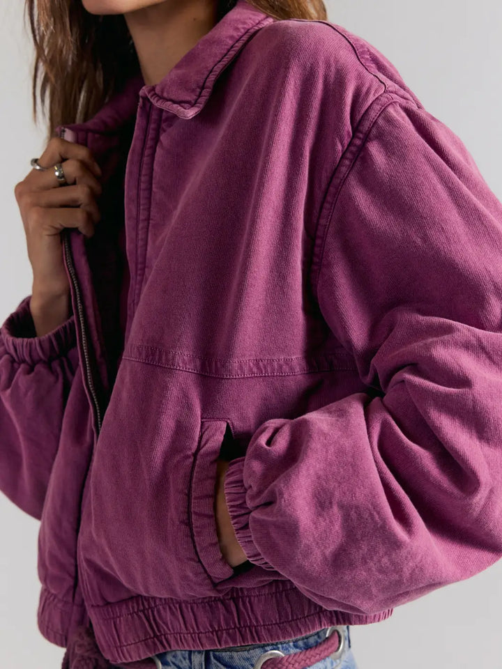 A close-up of a person wearing a dusty purple FP One Birdie Washed Jacket in Roan Rouge from Free People with elasticized cuffs and hem, paired with blue jeans against a light gray background | Twentyseven Toronto