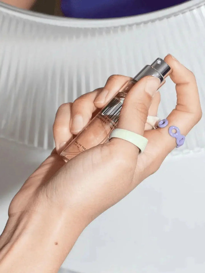 A person wearing colorful, chunky rings holds a small travel-sized spray vial of Dedcool Fragrance 05 "Spring" up toward a white, pleated lamp shade | Twentyseven Toronto