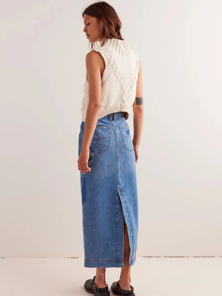 A rear view shows a model wearing the mid-wash Free People We The Free Francisco Denim Maxi Skirt - Head Over Heels featuring a high back slit, patch pockets, and a straight silhouette styled with a cream cable-knit vest | Twentyseven Toronto