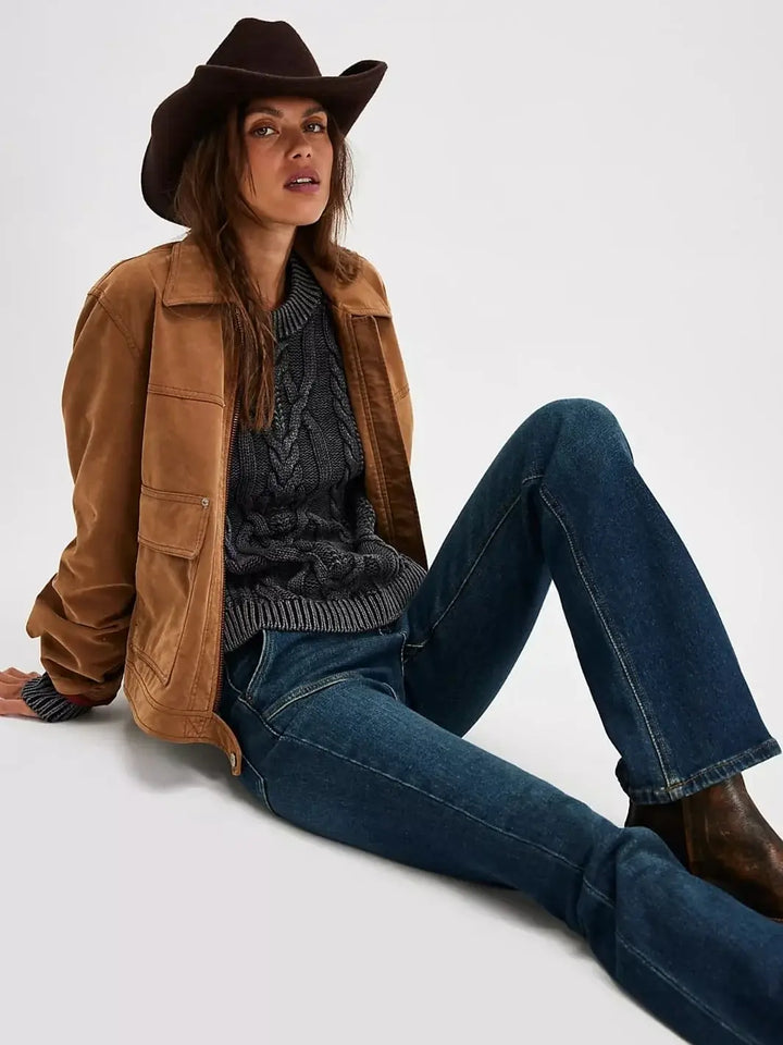 A model poses seated on the floor wearing the chunky Free People Frankie Cable Sweater in Black layered under a brown utility jacket and styled with flared blue jeans and a cowboy hat | Twentyseven Toronto