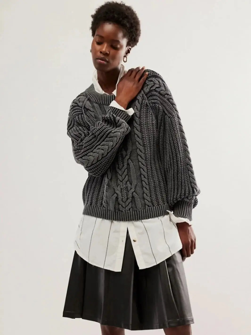 A full-length studio portrait captures a model wearing the relaxed-fit Free People Frankie Cable Sweater in Black styled over an oversized striped white shirt and a black leather pleated skirt | Twentyseven Toronto