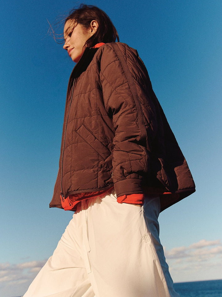 Free People Movement Pippa Packable Puffer in Cocoa styled with a red layer and white pants against a coastal backdrop | Twentyseven Toronto