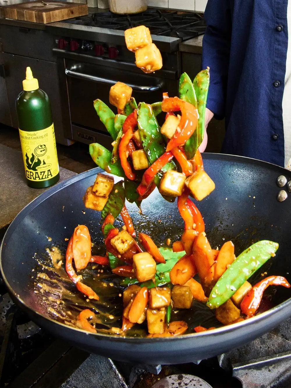 Stir-fry dish with vegetables in a wok on a stove, with a bottle of Graza 'Frizzle' High Heat Cooking Oil in the background | Twentyseven Toronto