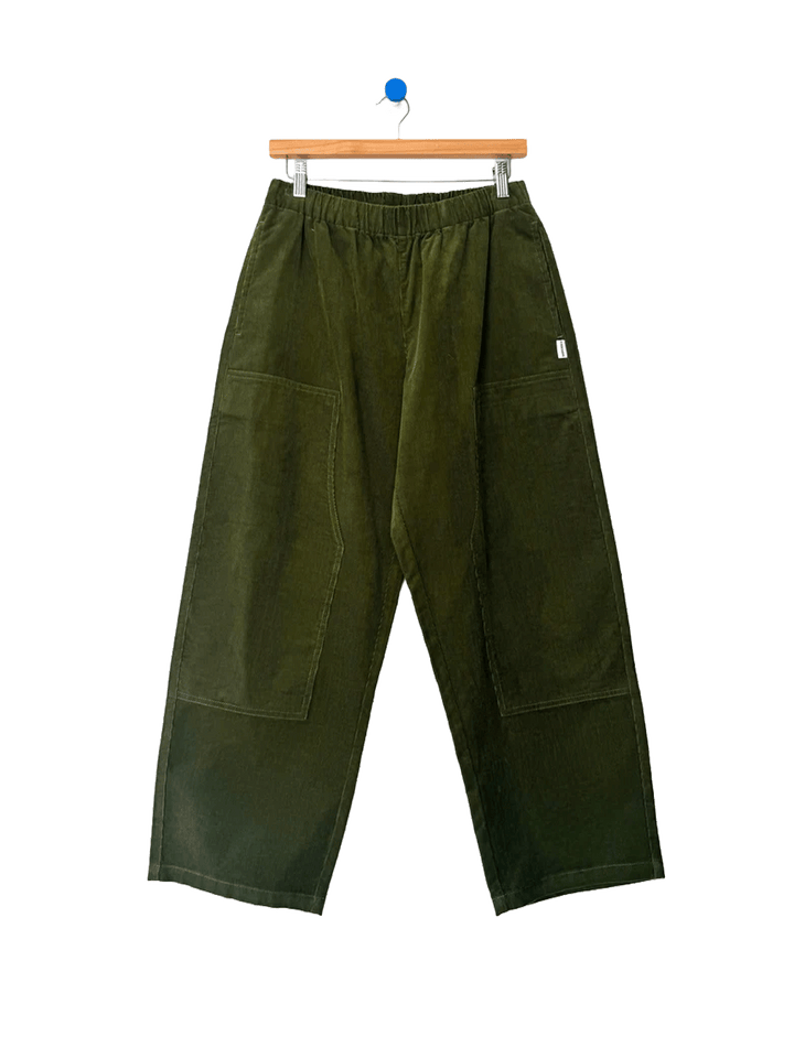 Le Bon Shoppe Corduroy Gardener's Pants in Bay Leaf on a wooden hanger against a white background, featuring an elastic waistband, oversized patch pockets and relaxed tapered legs | Twentyseven Toronto