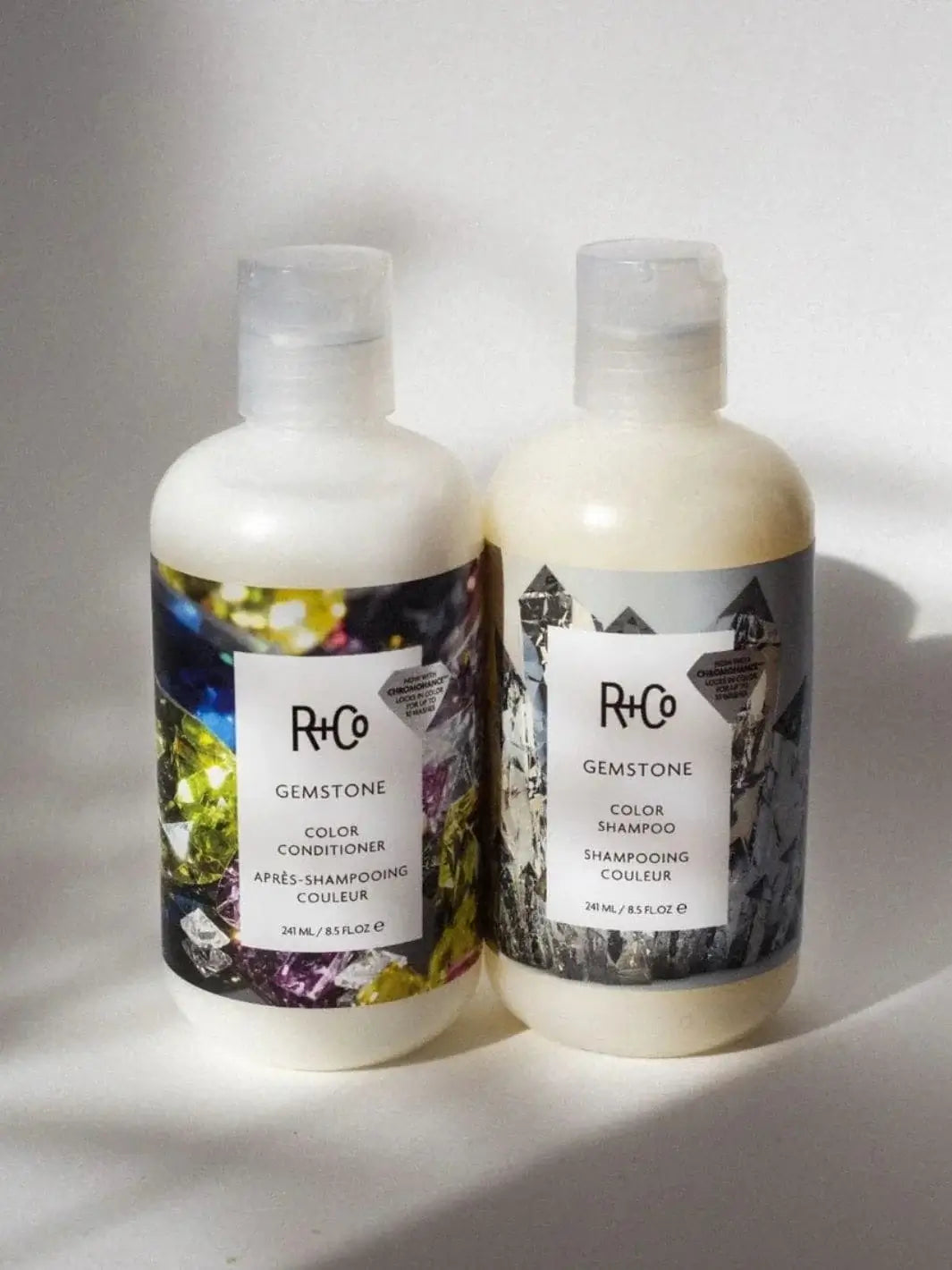 A bottle of R+Co GEMSTONE ChromoHance Color Shampoo stands upright next to its matching color conditioner under bright sunlight | Twentyseven Toronto