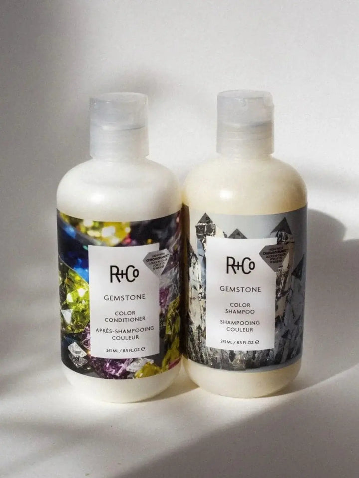 A bottle of R+Co GEMSTONE ChromoHance Color Shampoo stands upright next to its matching color conditioner under bright sunlight | Twentyseven Toronto