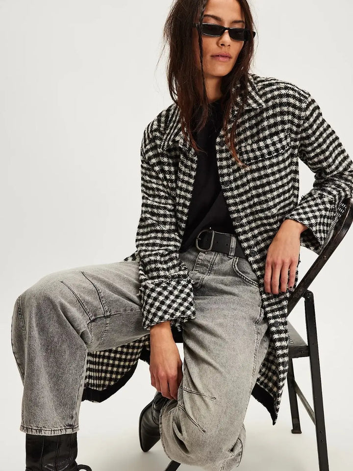 A model sits on a chair wearing the open Free People Get Around Houndstooth Shirt - Black + White styled over a black top and grey wash jeans | Twentyseven Toronto
