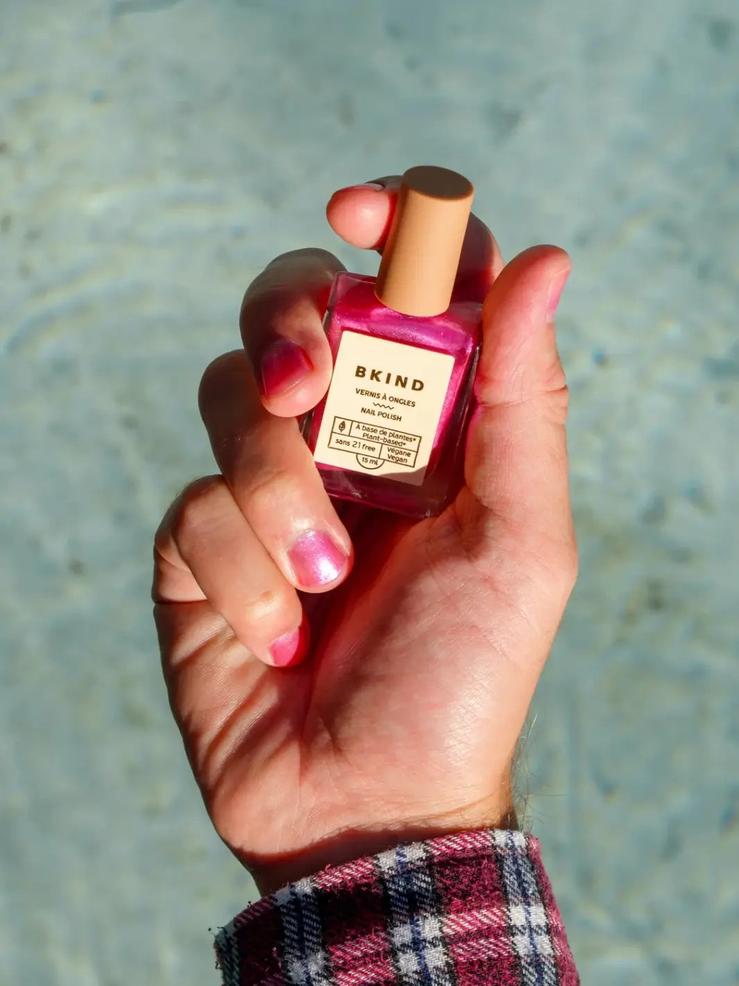 A close-up of a hand with a shimmering pink manicure holding a square glass bottle of BKIND Nail Polish in Britney against a backdrop of rippling pool water | Twentyseven Toronto