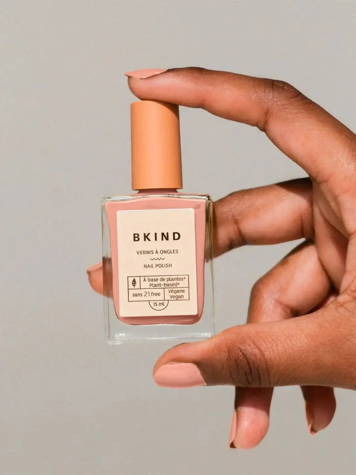 A close-up of a hand holding a square bottle of BKIND Nail Polish in Déjà Vu, displaying fingernails painted in the matching dusty rose pink shade | Twentyseven Toronto