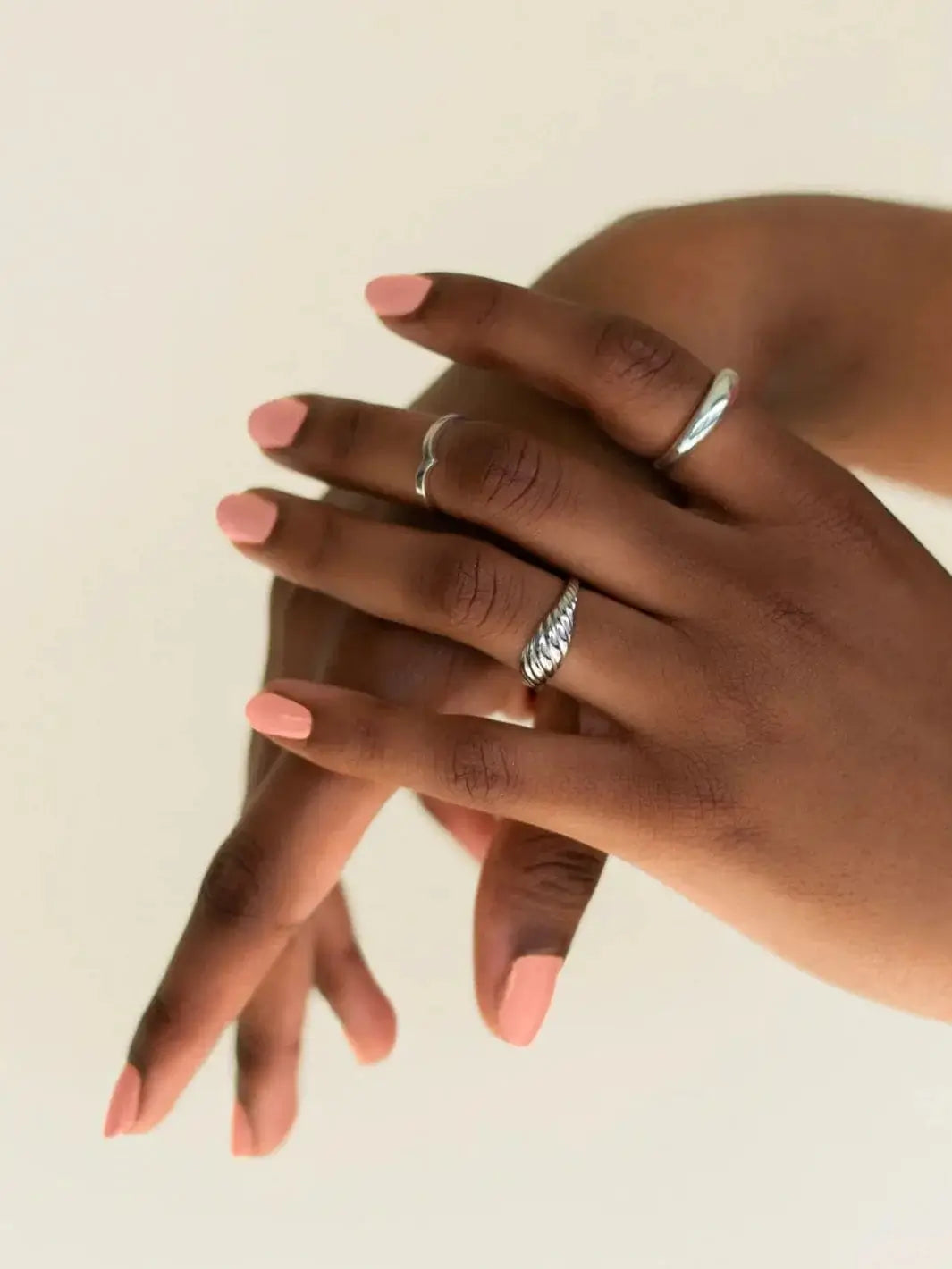 Hands adorned with silver rings showcase a neat, matte-finish pastel pink manicure featuring BKIND Nail Polish in Déjà Vu against an off-white background | Twentyseven Toronto