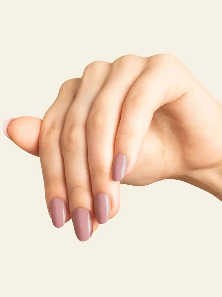 A hand showcases a neat manicure featuring the dusty mauve shade of BKIND Nail Polish in Wannabe against a cream background | Twentyseven Toronto