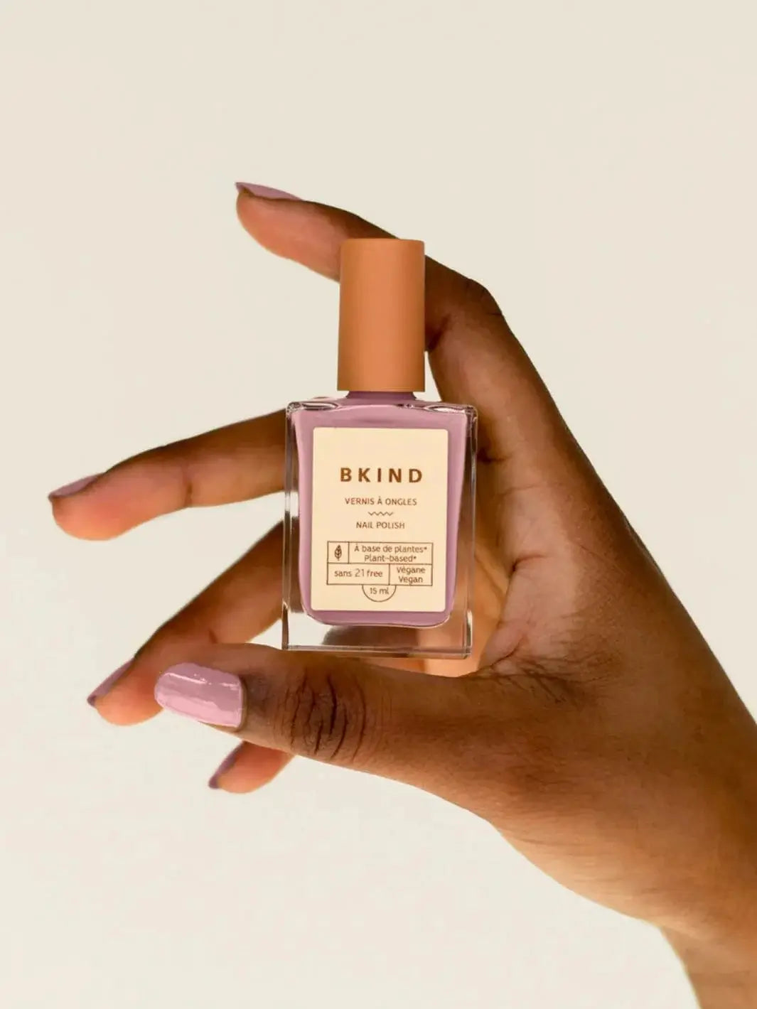 A hand holds up a square glass bottle of BKIND Nail Polish in Wannabe, displaying the matching mauve polish on the fingernails | Twentyseven Toronto