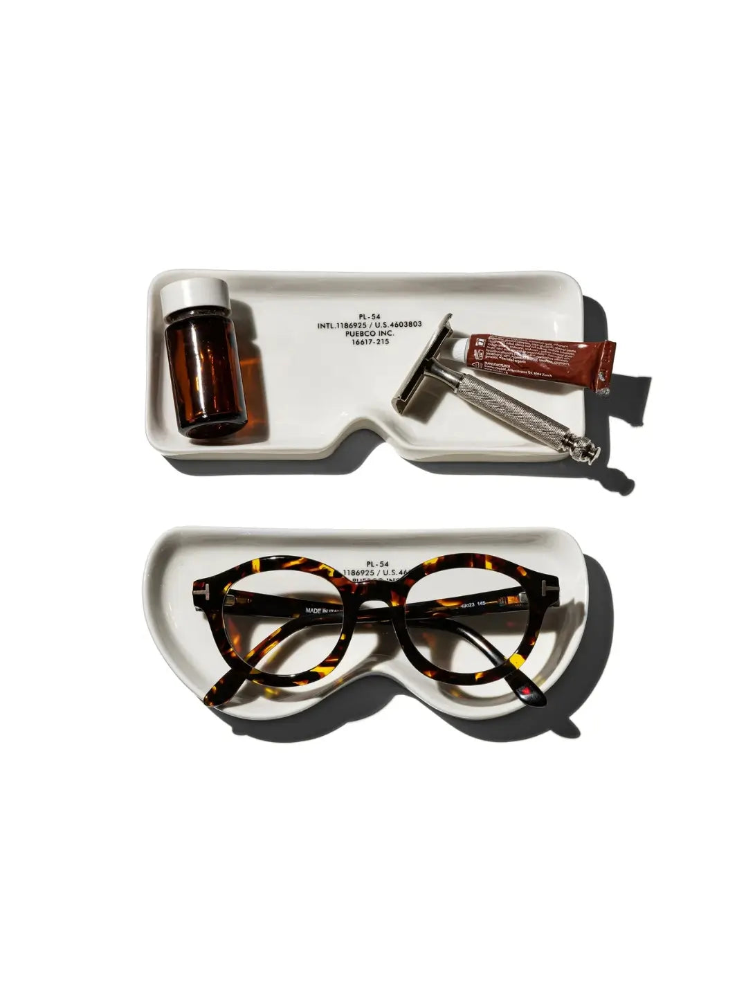 Two versions of the ceramic Glasses Tray - Round from Puebco are displayed against a white background, one holding grooming essentials like a razor and small bottle, and the other cradling a pair of tortoise shell eyeglasses | Twentyseven Toronto