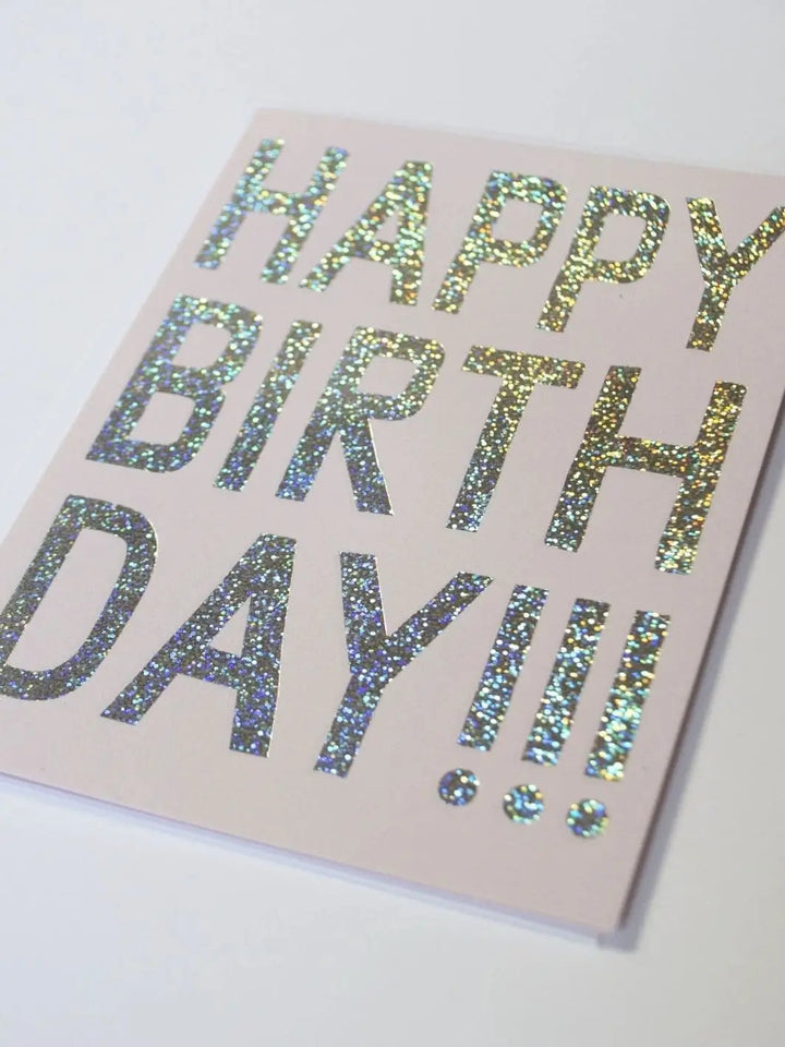 A close-up shot shows a light pink greeting card featuring the words "HAPPY BIRTHDAY!!!" rendered in bold, shimmering silver glitter, showcasing the Banquet Workshop Glitter Hologram Foil Birthday Card design | Twentyseven Toronto