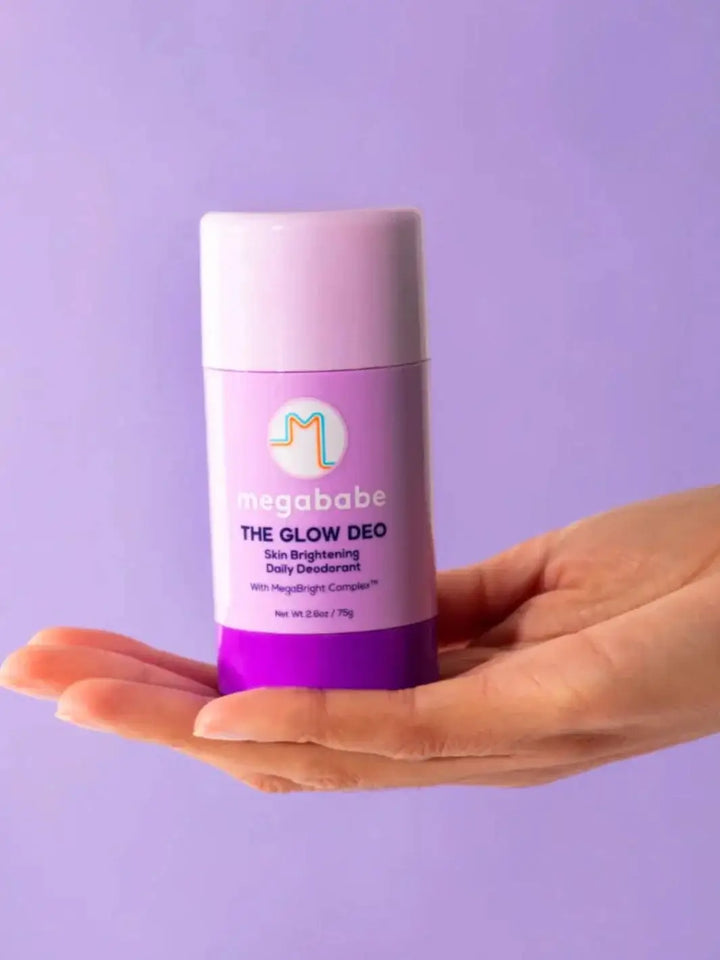 A hand holds up a purple container of Megababe The Glow Deo centered against a solid lavender background | Twentyseven Toronto