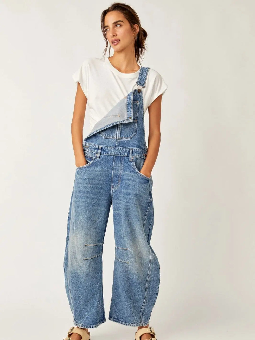A front studio perspective captures a model posing with hands in pockets, wearing the Free People Good Luck Overall in Ultra Light Beam with one strap loosely unbuttoned | Twentyseven Toronto