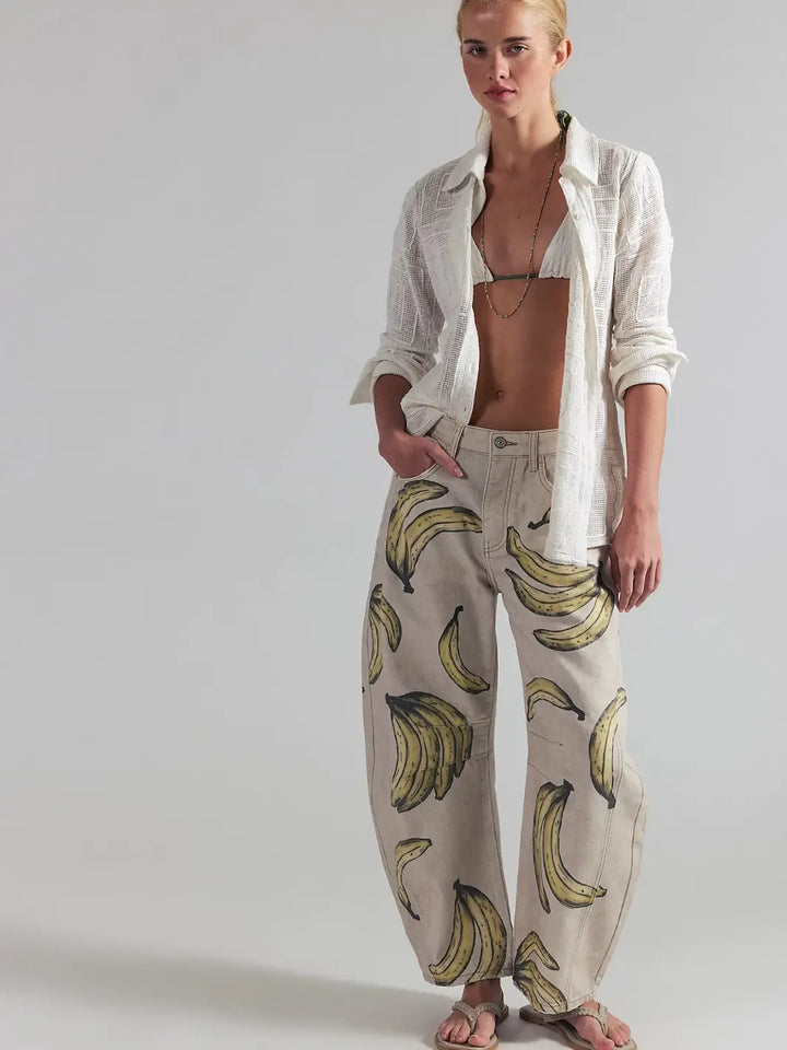 A full-length front view of a model posing in an open white button-down shirt, bikini top, and the Free People Good Luck Printed Barrel Jeans in Go Bananas | Twentyseven Toronto