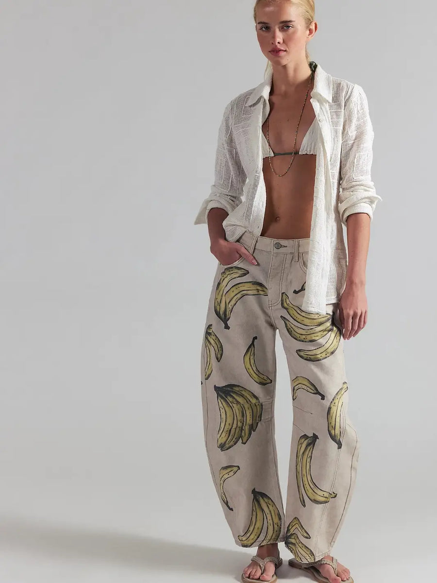 A full-length front view of a model posing in an open white button-down shirt, bikini top, and the Free People Good Luck Printed Barrel Jeans in Go Bananas | Twentyseven Toronto