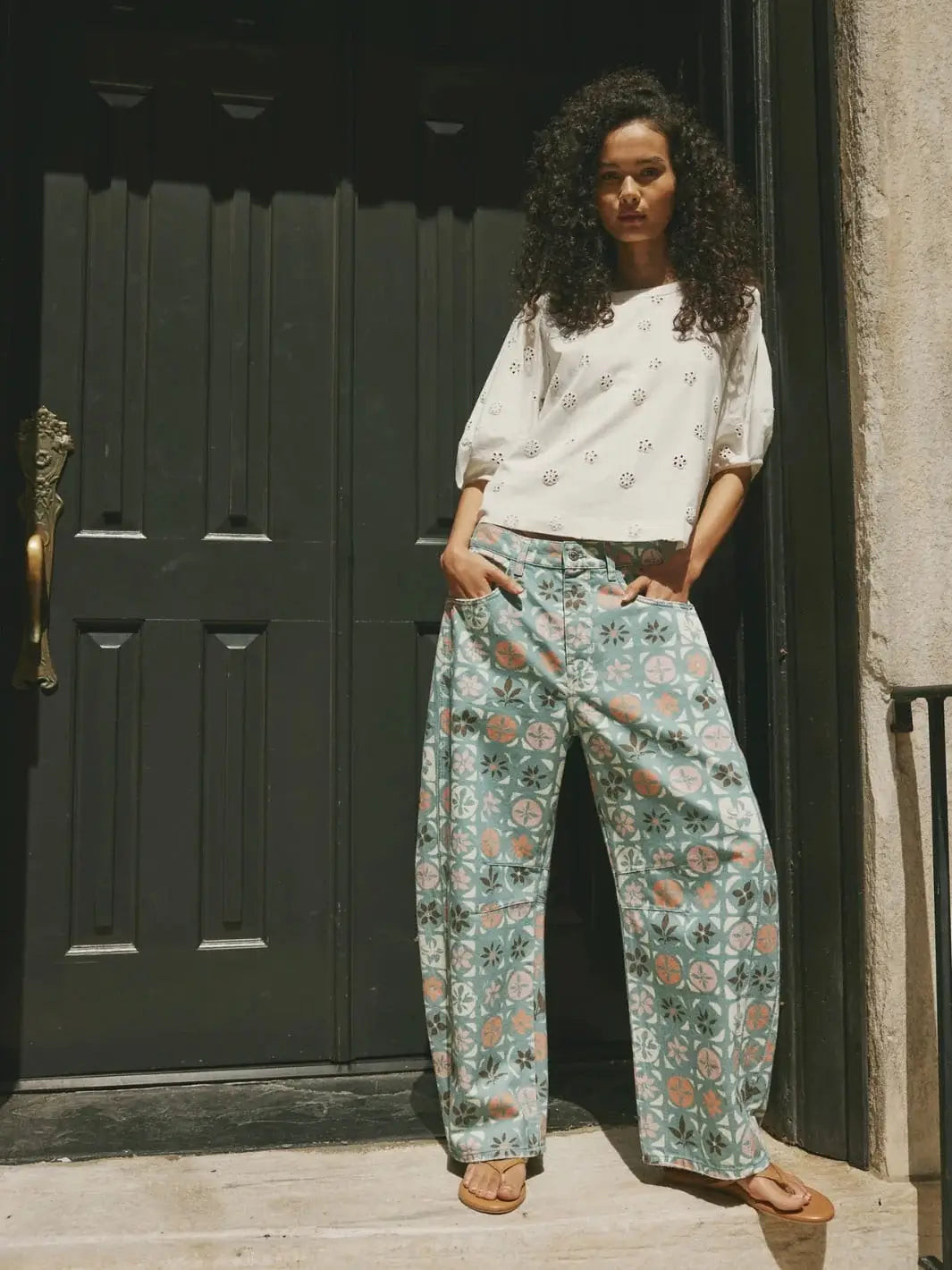 An outdoor shot captures a model standing in front of a dark door while wearing the patterned Free People Good Luck Printed Barrel Jeans in Jade Combo Ditsy Block paired with an eyelet-detailed white short-sleeve top | Twentyseven Toronto