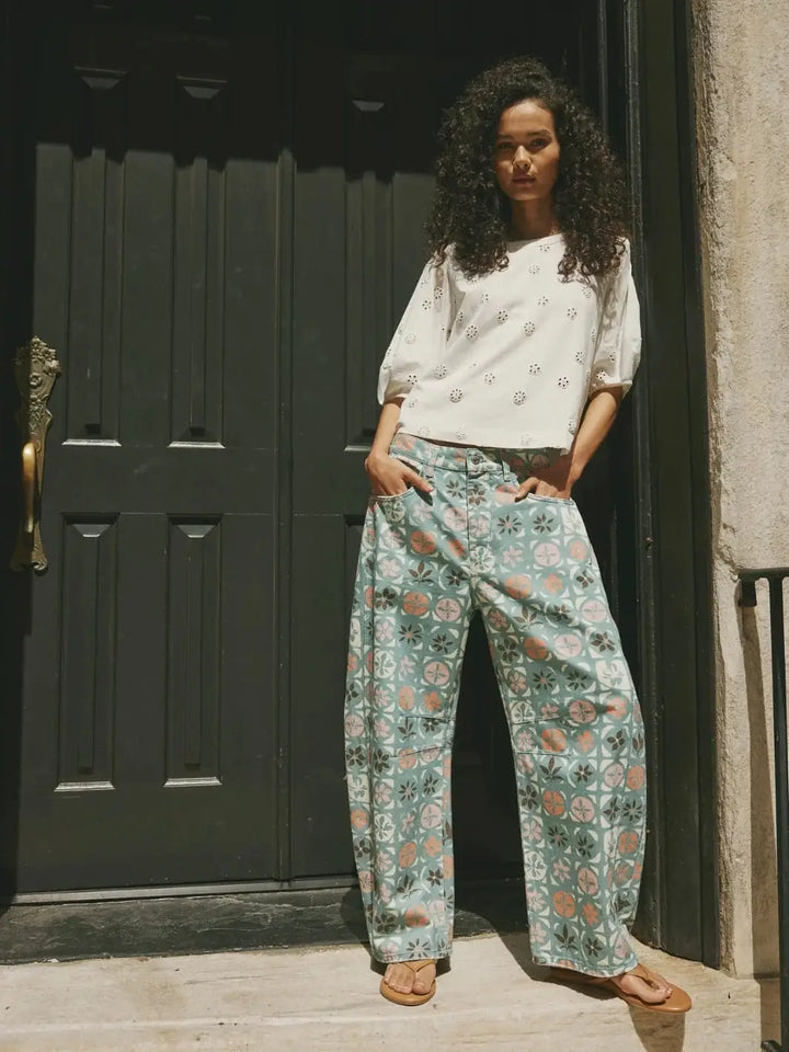 An outdoor shot captures a model standing in front of a dark door while wearing the patterned Free People Good Luck Printed Barrel Jeans in Jade Combo Ditsy Block paired with an eyelet-detailed white short-sleeve top | Twentyseven Toronto