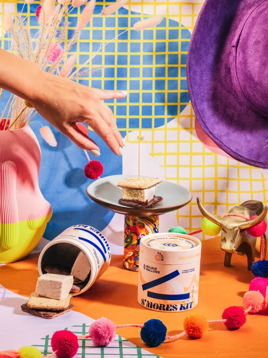 A hand reaches toward an assembled s'more on a small pedestal plate, surrounded by colourful retro decorations and cylindrical packaging from the Mojave Mallows Gourmet S'mores Kit | Twentyseven Toronto