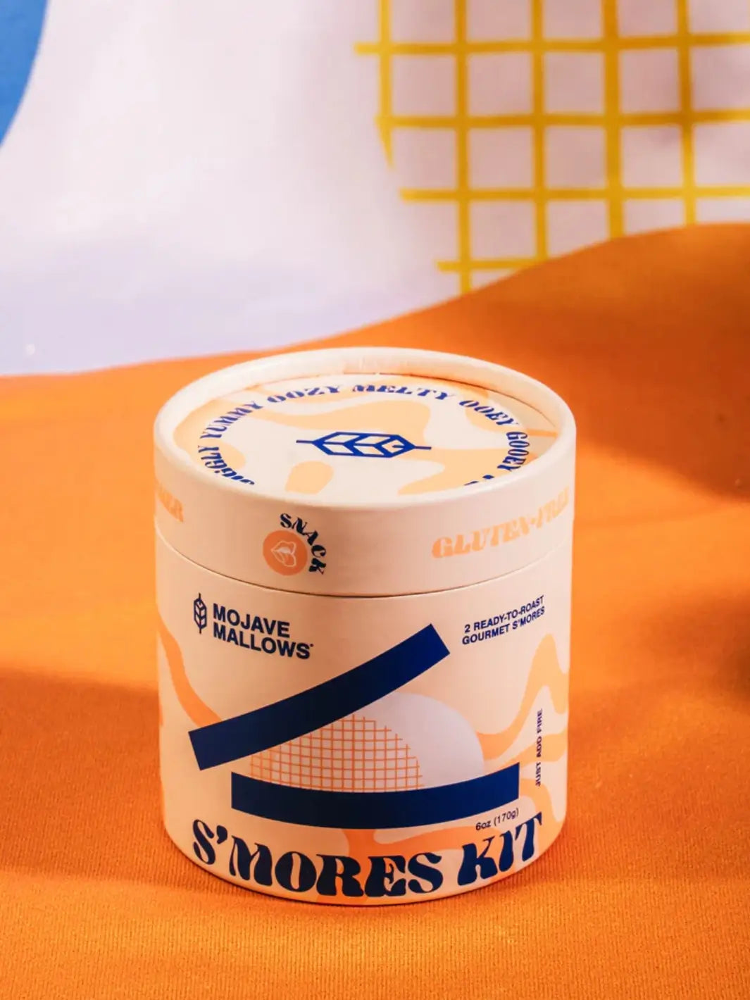 A close-up of the cylindrical tub packaging of the Mojave Mallows Gourmet S'mores Kit resting on an orange fabric surface | Twentyseven Toronto