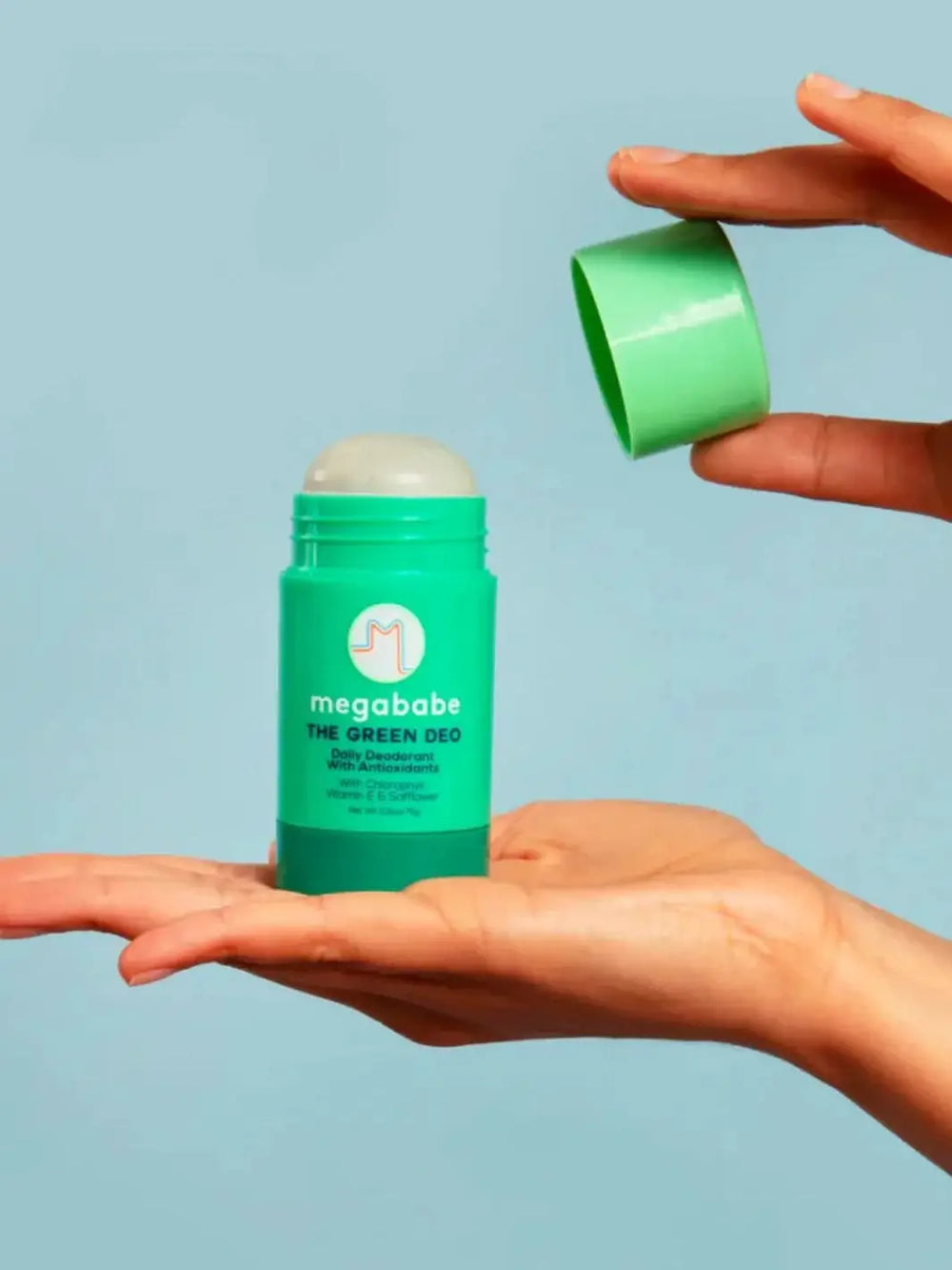 A person holds an opened green stick of Megababe The Green Deo in their palm against a solid light blue background while lifting off the cap | Twentyseven Toronto