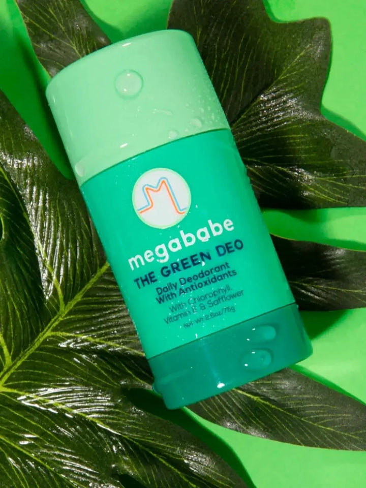 A green container of Megababe The Green Deo lies flat on top of large, wet green leaves to highlight its natural antioxidant formula | Twentyseven Toronto