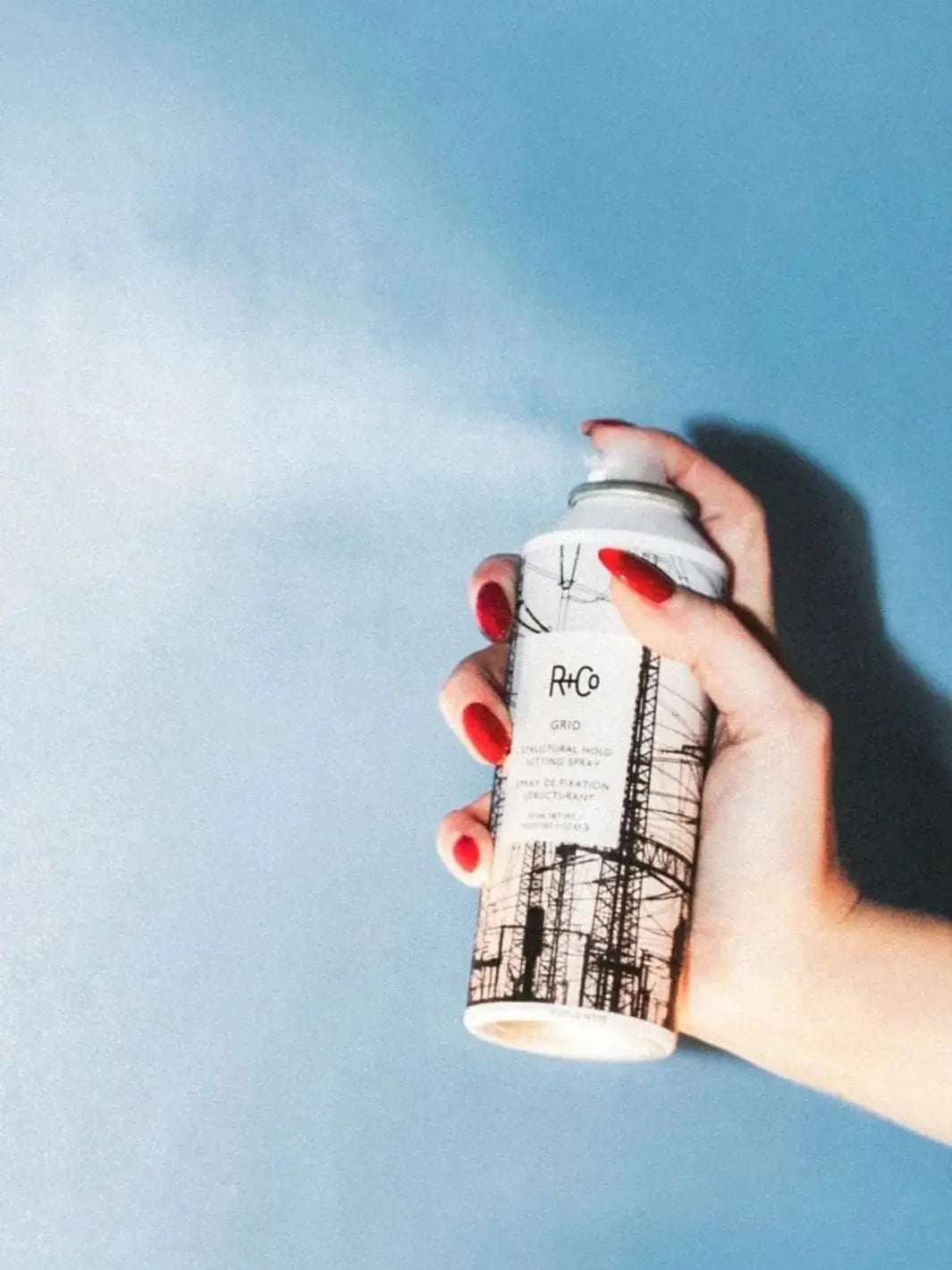 A hand with bright red fingernails presses the nozzle of an aerosol can, releasing a fine mist of R+Co GRID Structural Hold Setting Spray against a blue background | Twentyseven Toronto
