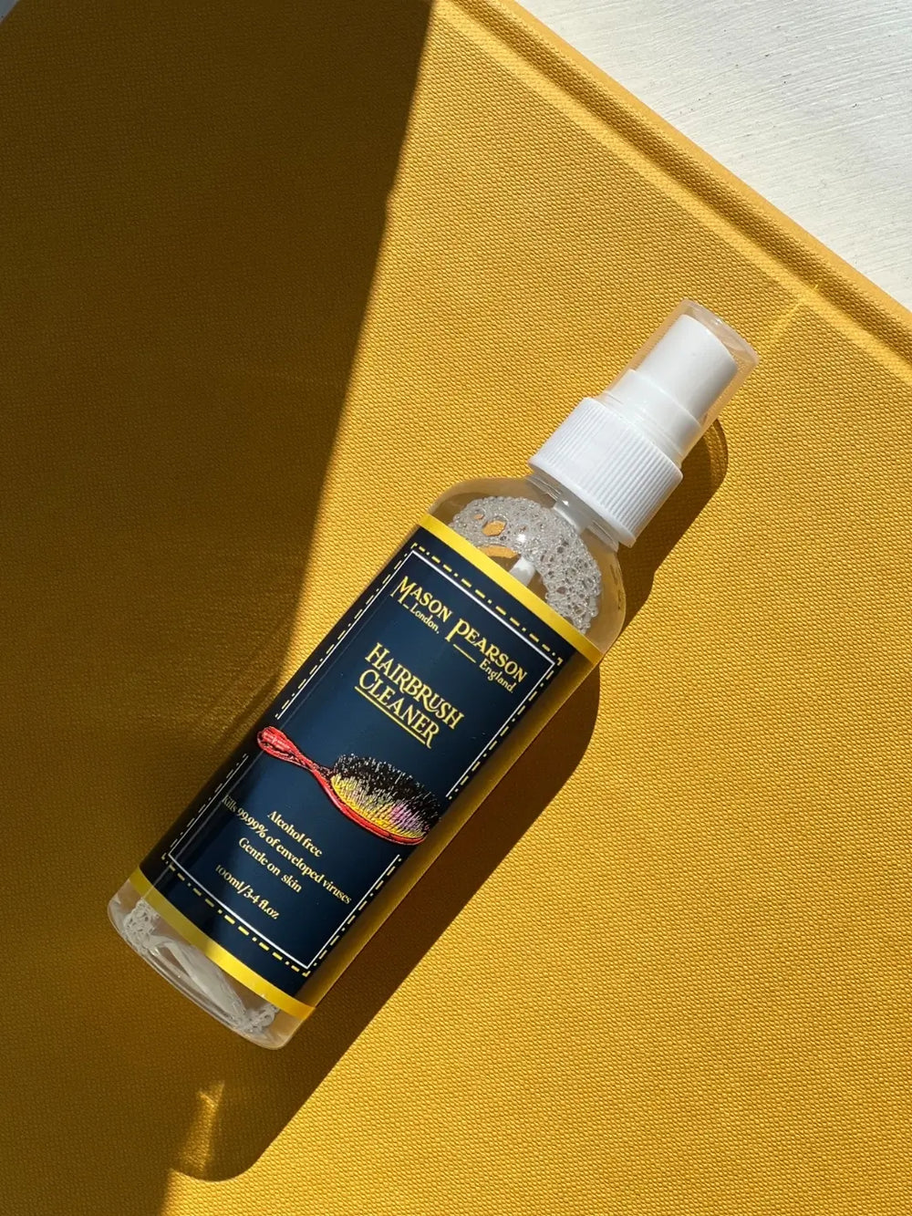 A clear spray bottle featuring a dark blue and yellow label is placed diagonally across a textured mustard yellow surface to highlight the Mason Pearson Hairbrush Cleaner | Twentyseven Toronto