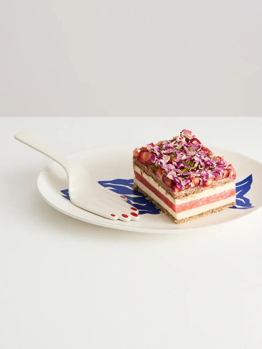 Layered dessert with pink and white flowers on a decorative plate beside a hand cake server shaped as a hand with red nail polish with Maison Balzac against a white background | Twentyseven Toronto