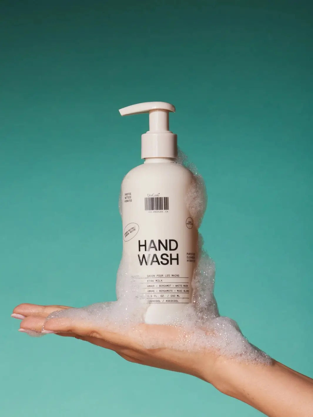 A hand holding a cream-coloured pump bottle of Dedcool Hand Wash Refill Xtra Milk surrounded by foamy soap suds against a green background | Twentyseven