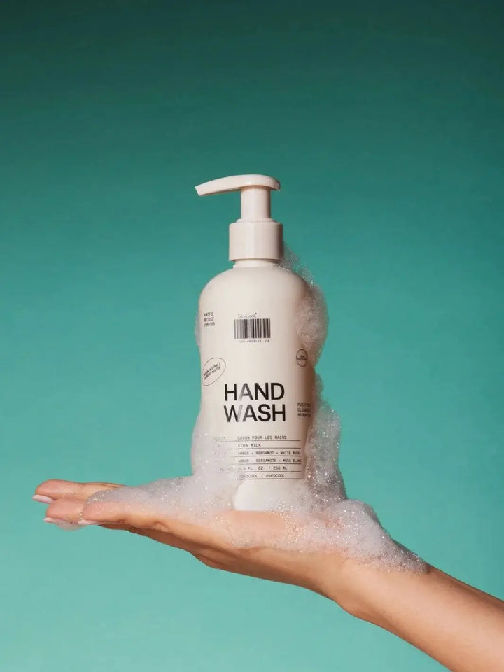 A hand holds up a bottle of Dedcool Hand Wash 01 "Taunt" covered in white, foamy soap bubbles against a solid teal background | Twentyseven Toronto