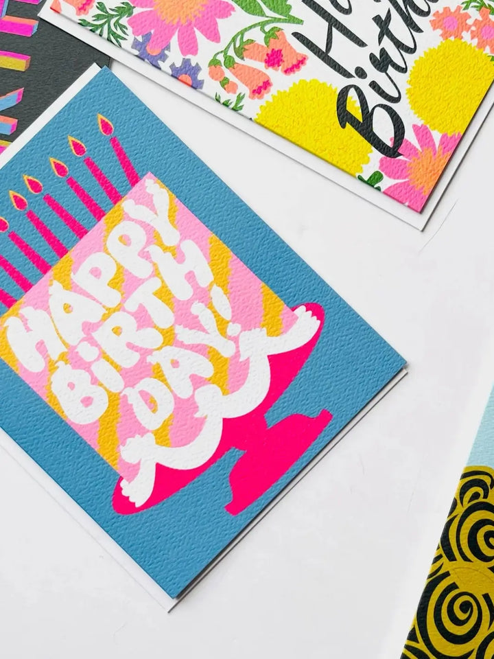 A close-up, angled view highlights the textured cardstock of the Banquet Workshop Happy Birthday Pink Cake Dreams Note Card as it lies next to a bright floral card | Twentyseven